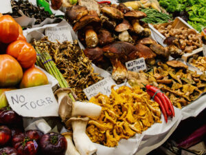 Colorful market stall displaying a variety of fresh mushrooms, including chanterelles, porcini, and king oyster mushrooms, alongside tomatoes, asparagus, red chili peppers, and other vegetables, with handwritten “No Tocar” signs in Spanish indicating not to touch the produce.