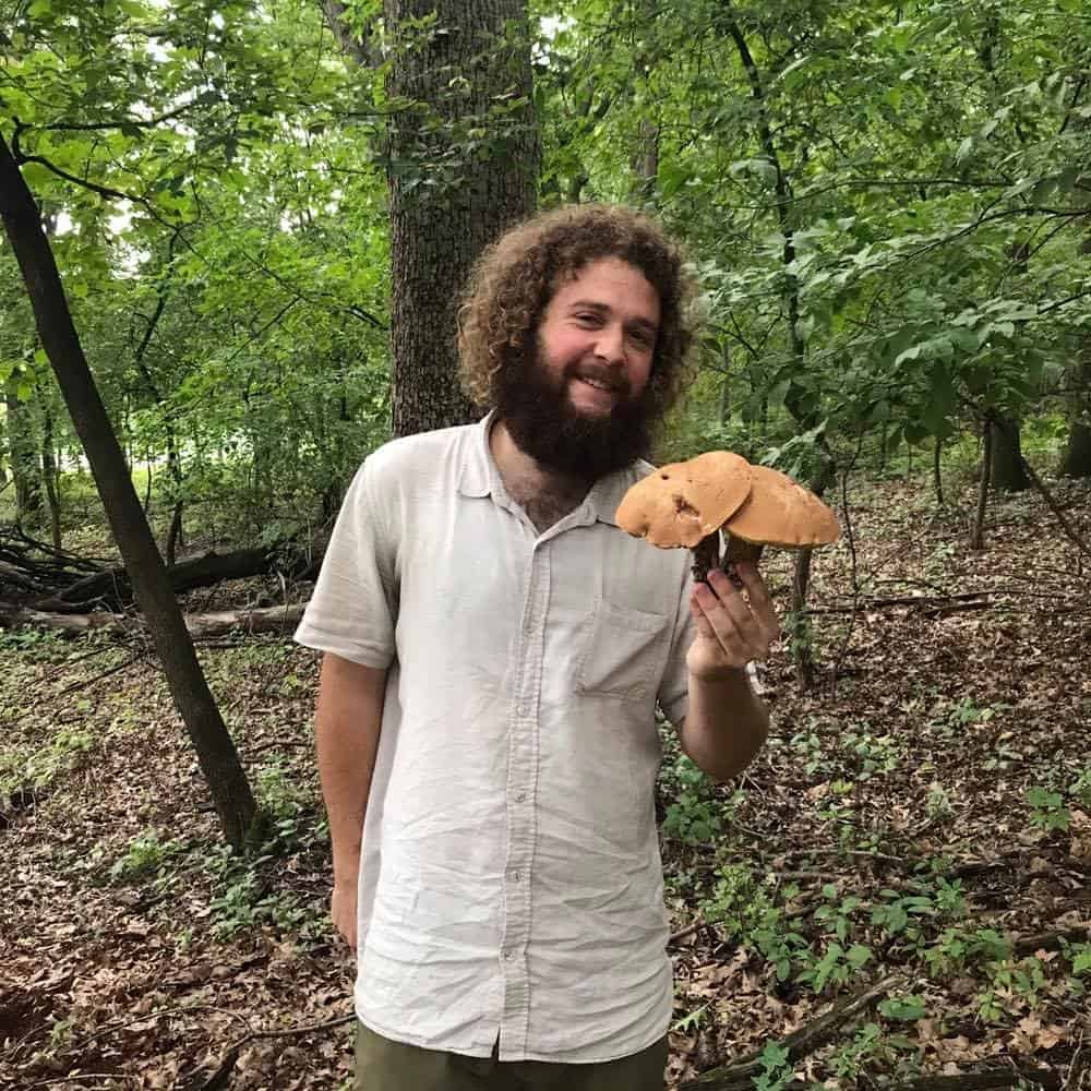 Colin Peters stands in a forest clearing holding a cluster of large, tan-capped Lanmaoa mushrooms. He smiles at the camera, wearing a light short-sleeved button-up shirt, surrounded by leafy undergrowth and tall trees.