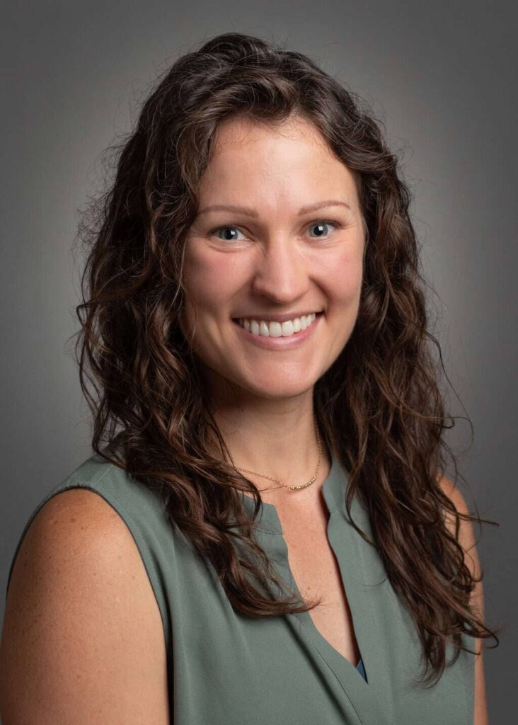 Professional headshot of Kathleen Thompson, smiling against a neutral gray background. She has long, wavy brown hair and wears a sleeveless green blouse with a delicate necklace.