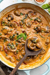 A creamy mushroom dish simmering in a skillet with sliced mushrooms, fresh basil, sun-dried tomatoes, and a wooden spoon scooping up a serving.