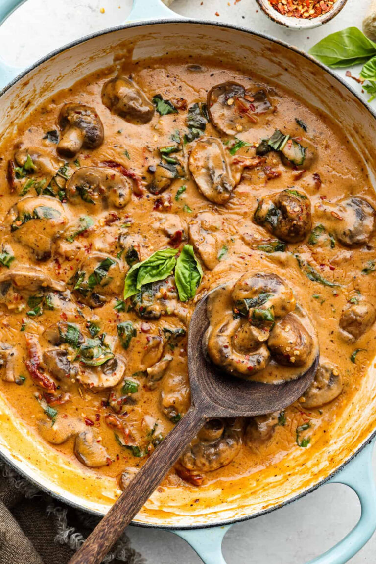 A creamy mushroom dish simmering in a skillet with sliced mushrooms, fresh basil, sun-dried tomatoes, and a wooden spoon scooping up a serving.