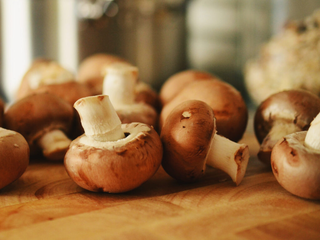 Cremini mushrooms (baby bellas) arranged on a wooden cutting board in a warmly lit kitchen, with a blurred background showing metal pots and other kitchen items.