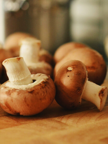 Cremini mushrooms (baby bellas) arranged on a wooden cutting board in a warmly lit kitchen, with a blurred background showing metal pots and other kitchen items.