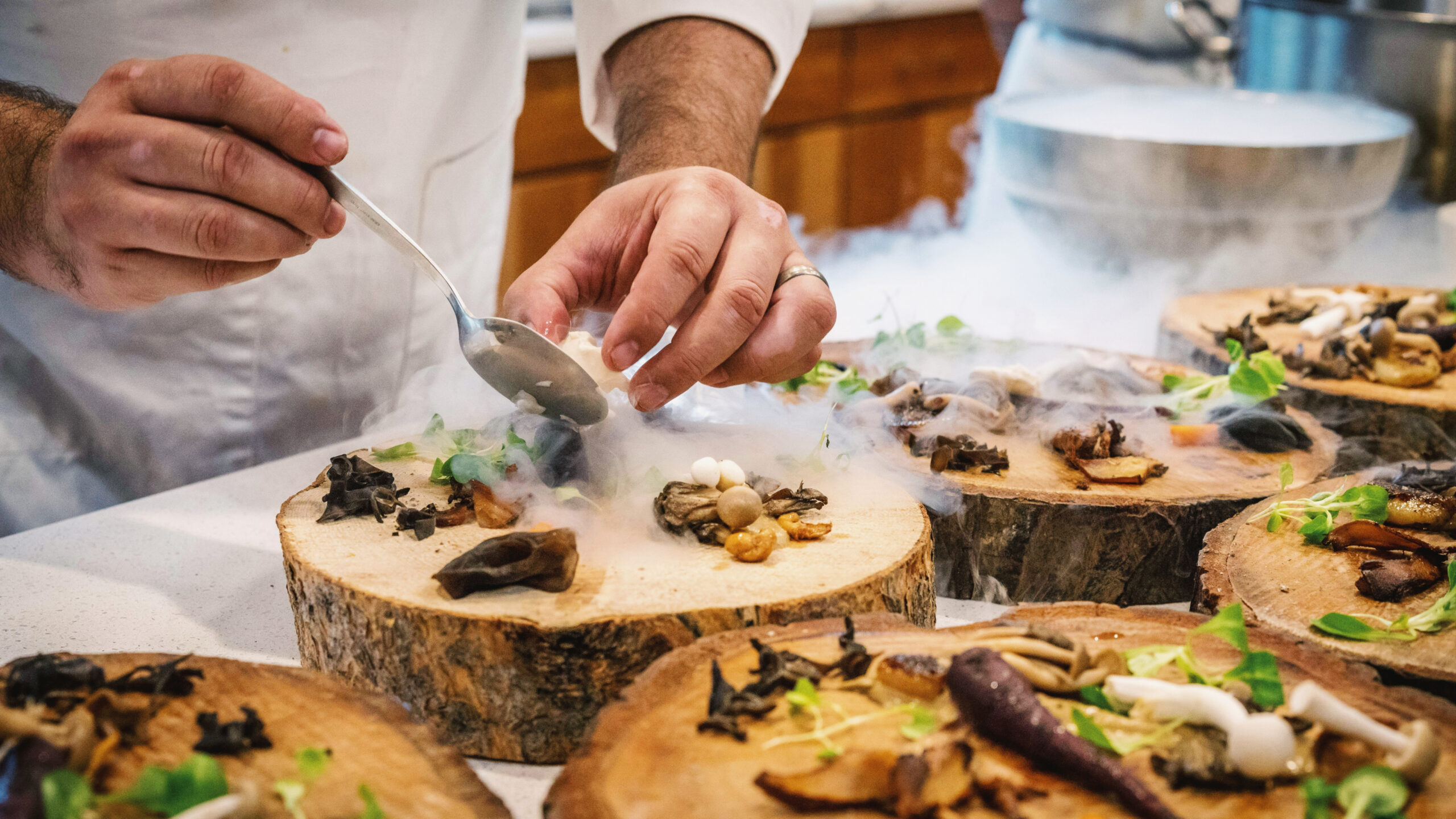 A chef carefully plates a vegetable and mushroom dish on a rustic tree slab, with wisps of smoke rising from the presentation, creating a dramatic and artistic culinary display.
