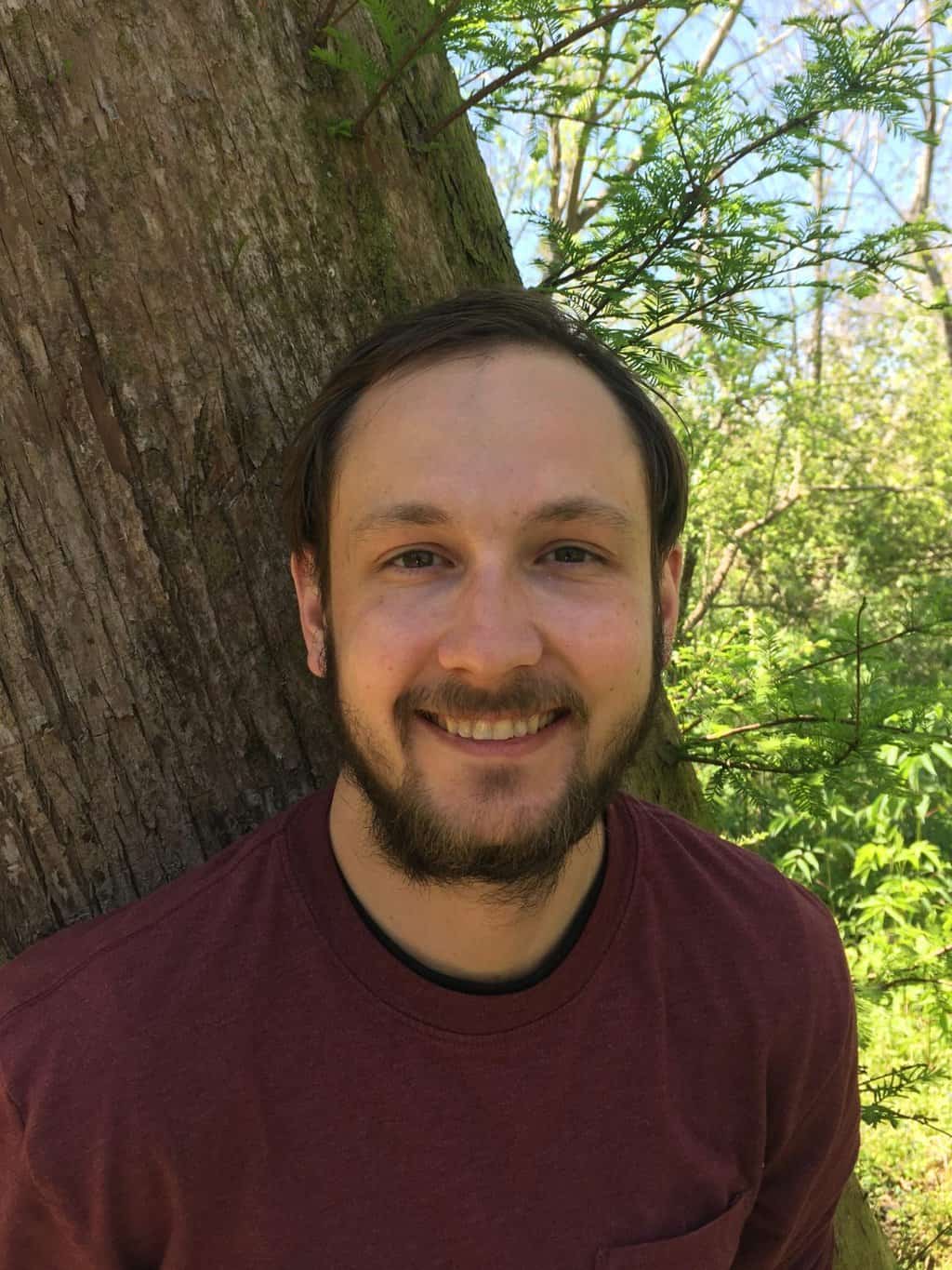 A man with short brown hair and a beard smiles while leaning against a tree in a wooded area. He is wearing a maroon shirt, and sunlight filters through the green foliage in the background.