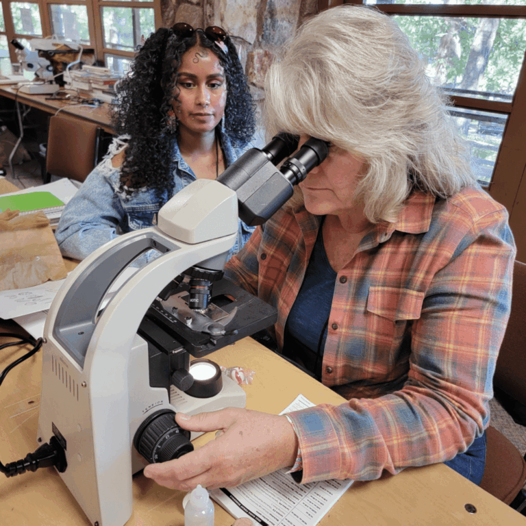 An instructor uses a microscope while a student looks on during a NAMA microscopy lab focused on fungal spores.