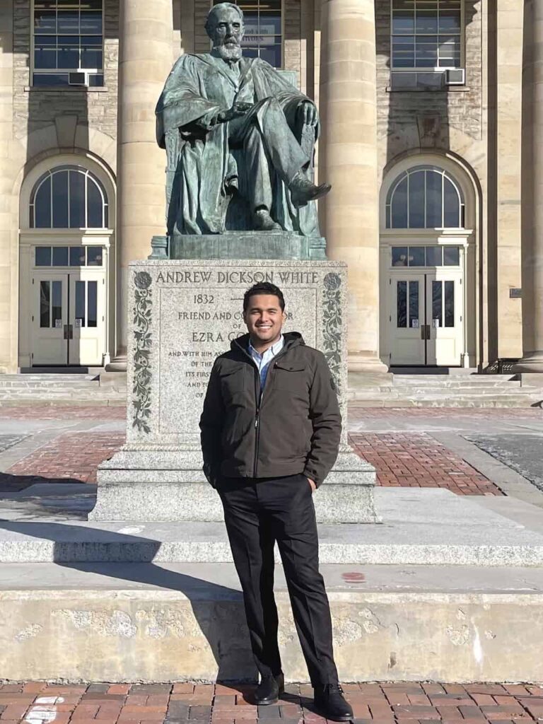 A man standing and smiling in front of the Andrew Dickson White statue on a university campus. The large seated bronze statue is mounted on a granite pedestal with engraved text and flanked by tall columns and arched windows.