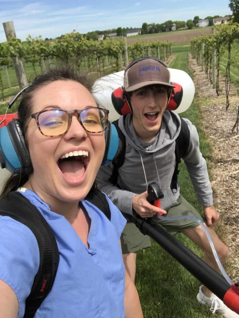 Two vineyard workers smiling and posing for a selfie, wearing hearing protection and backpacks for spraying. Rows of grapevines and an open field stretch out behind them under a bright sky.